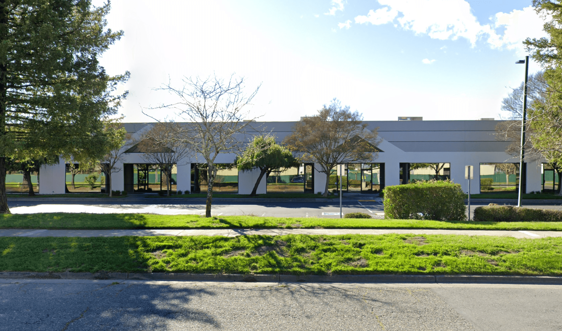 Modern white commercial building with large glass windows, landscaped with green grass and trees.